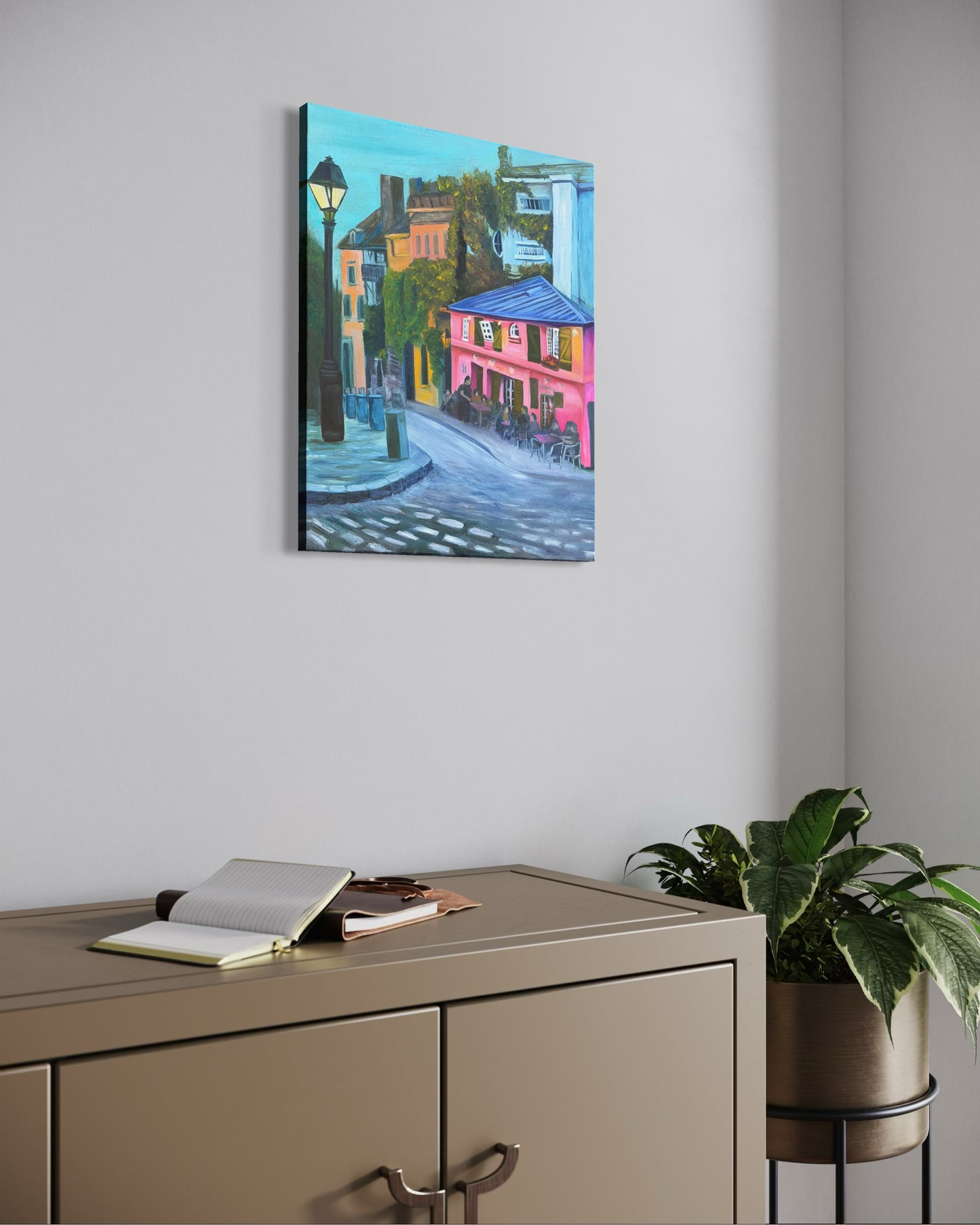 Colorful oil painting of a street scene with a pink cafe hanging on a wall above a cabinet with a plant.