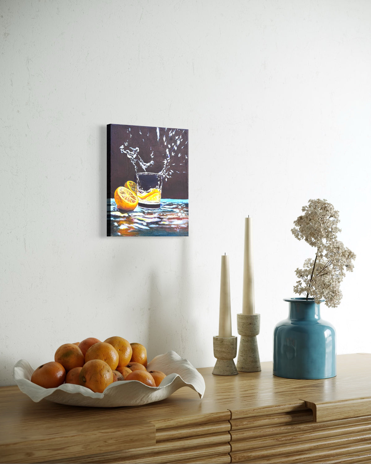 Canvas oil painting of a cup of water splashing out from an lemon slice dropped in and two halves beside the cup. Hung on a wall above a wooden table with decorative items.