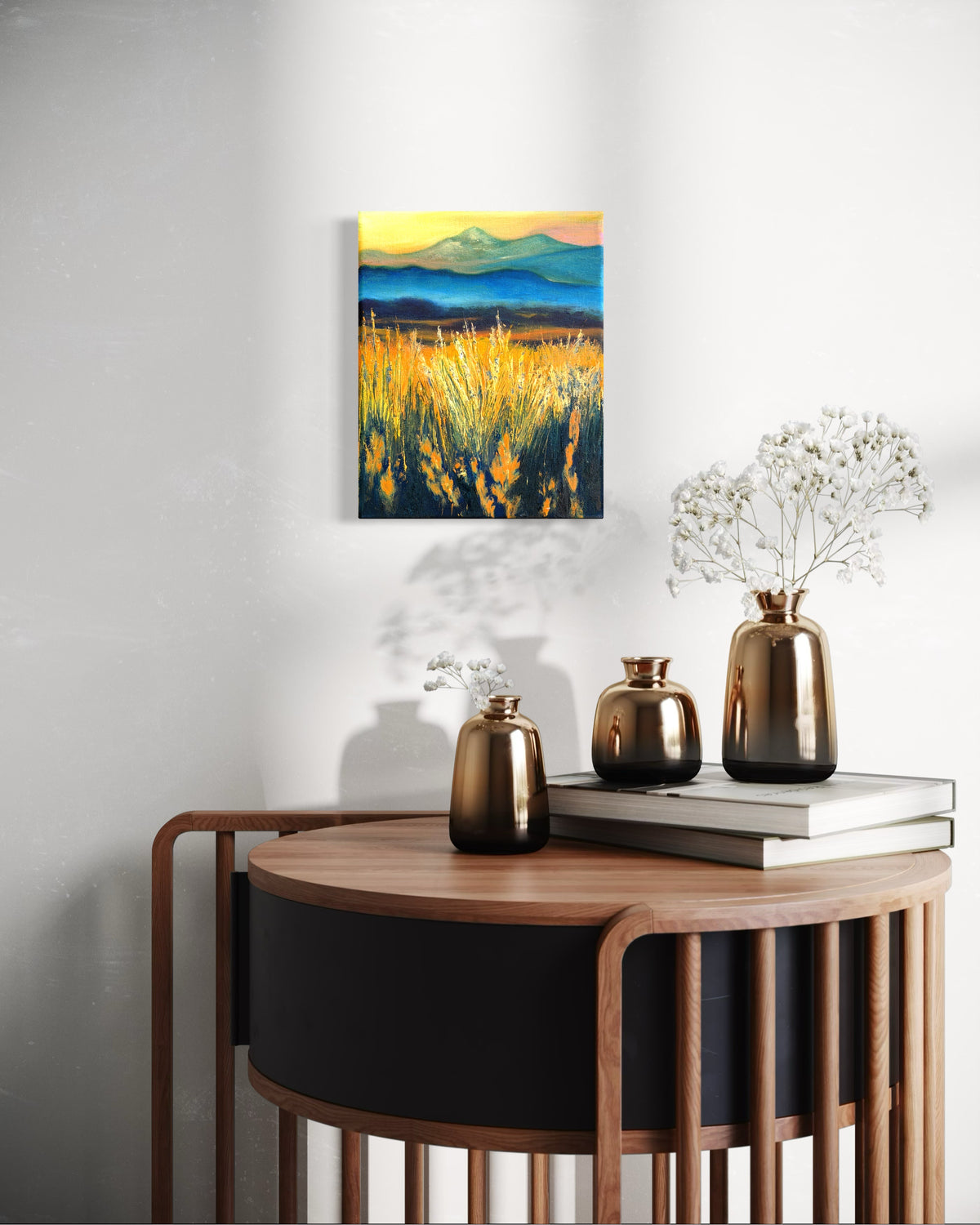 Decorative setting with a round wooden table and vases. An oil painting of a faded mountain landscape with tall grass glowing from the sun hangs on the wall.