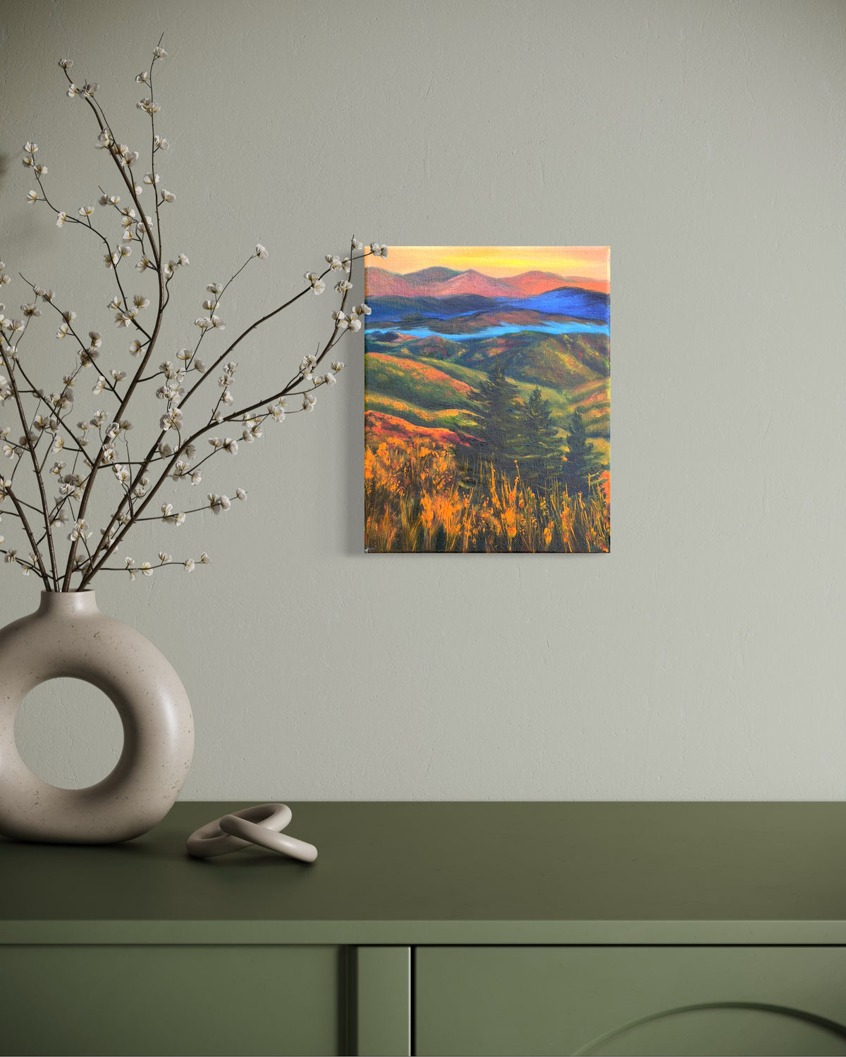 Oil painting of a landscape with rolling hills, trees, and a river in-between the mountains at sunset hanging on a wall above a green cabinet with decorative elements.