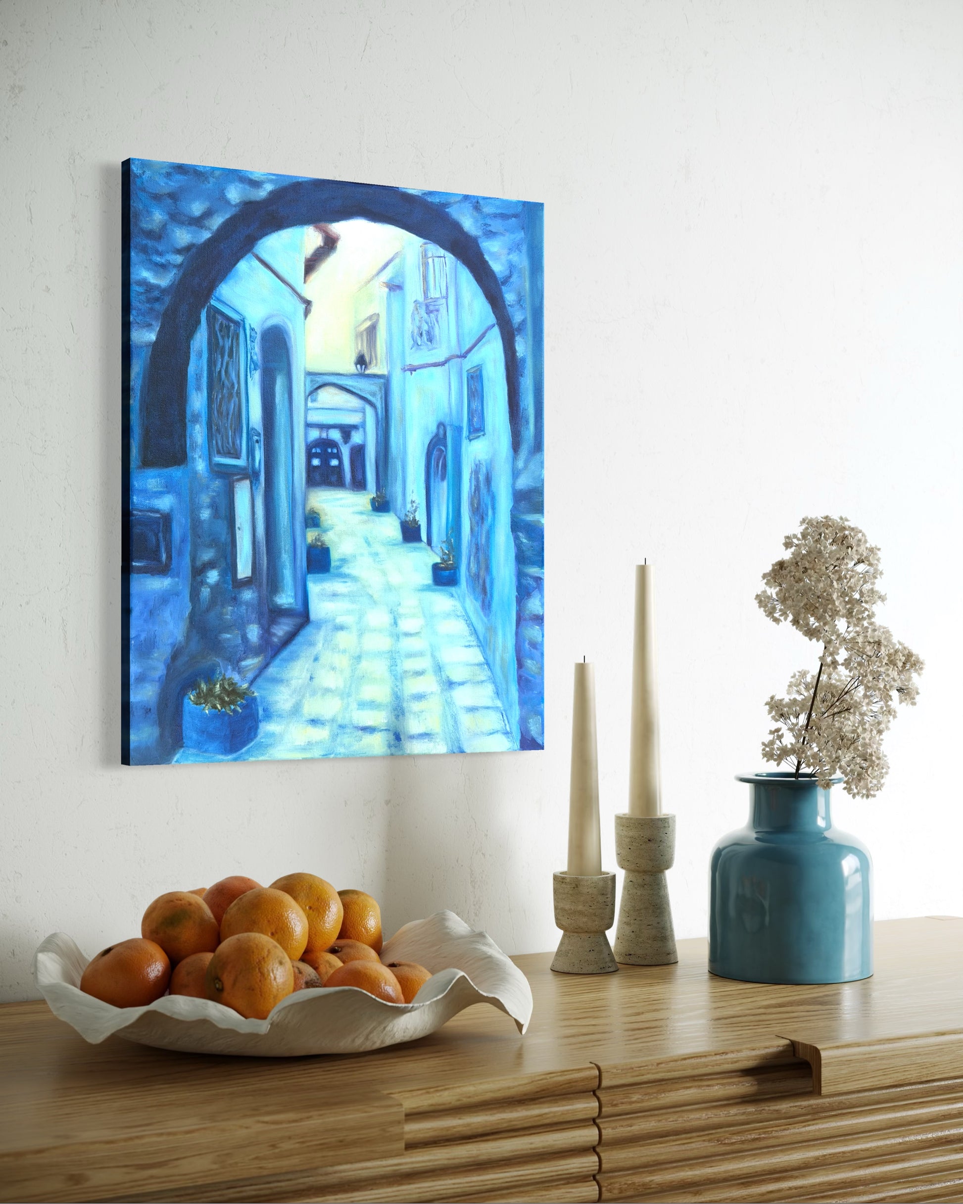 Decorative oil painting of a blue alleyway on a wall above a wooden shelf with a bowl of oranges, candles, and a vase.