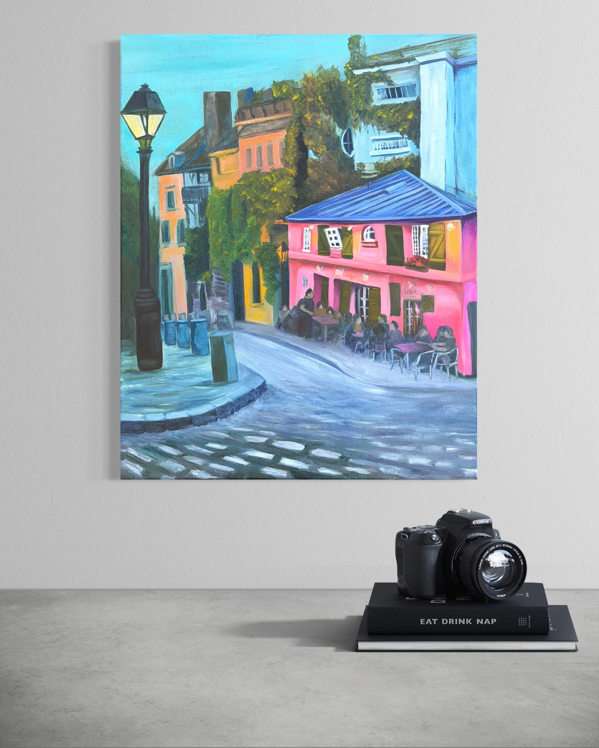 Colorful oil painting of a street scene with a pink cafe hanging on a wall with a camera below.