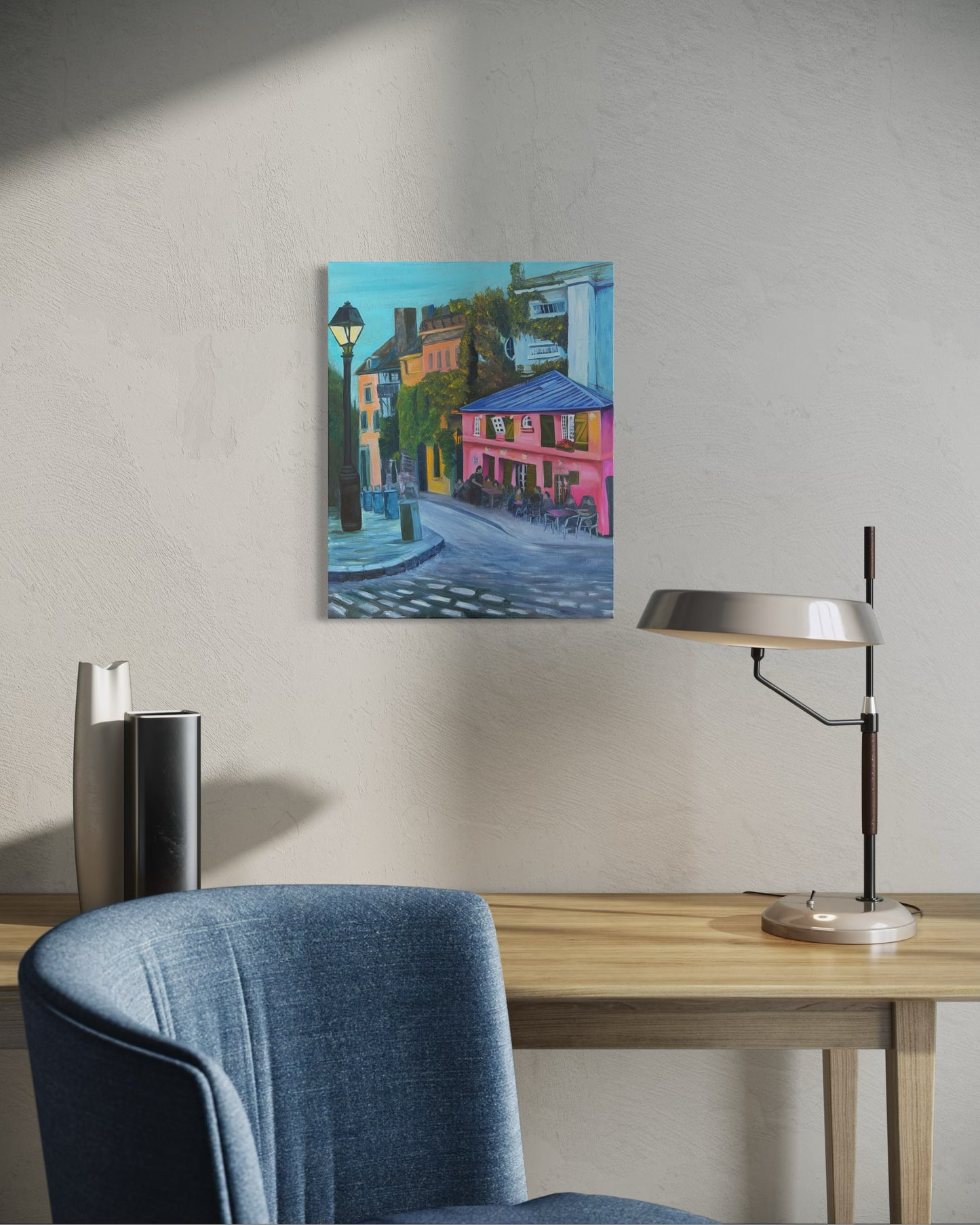 Colorful oil painting of a street scene on a wall above a desk with a chair and lamp.