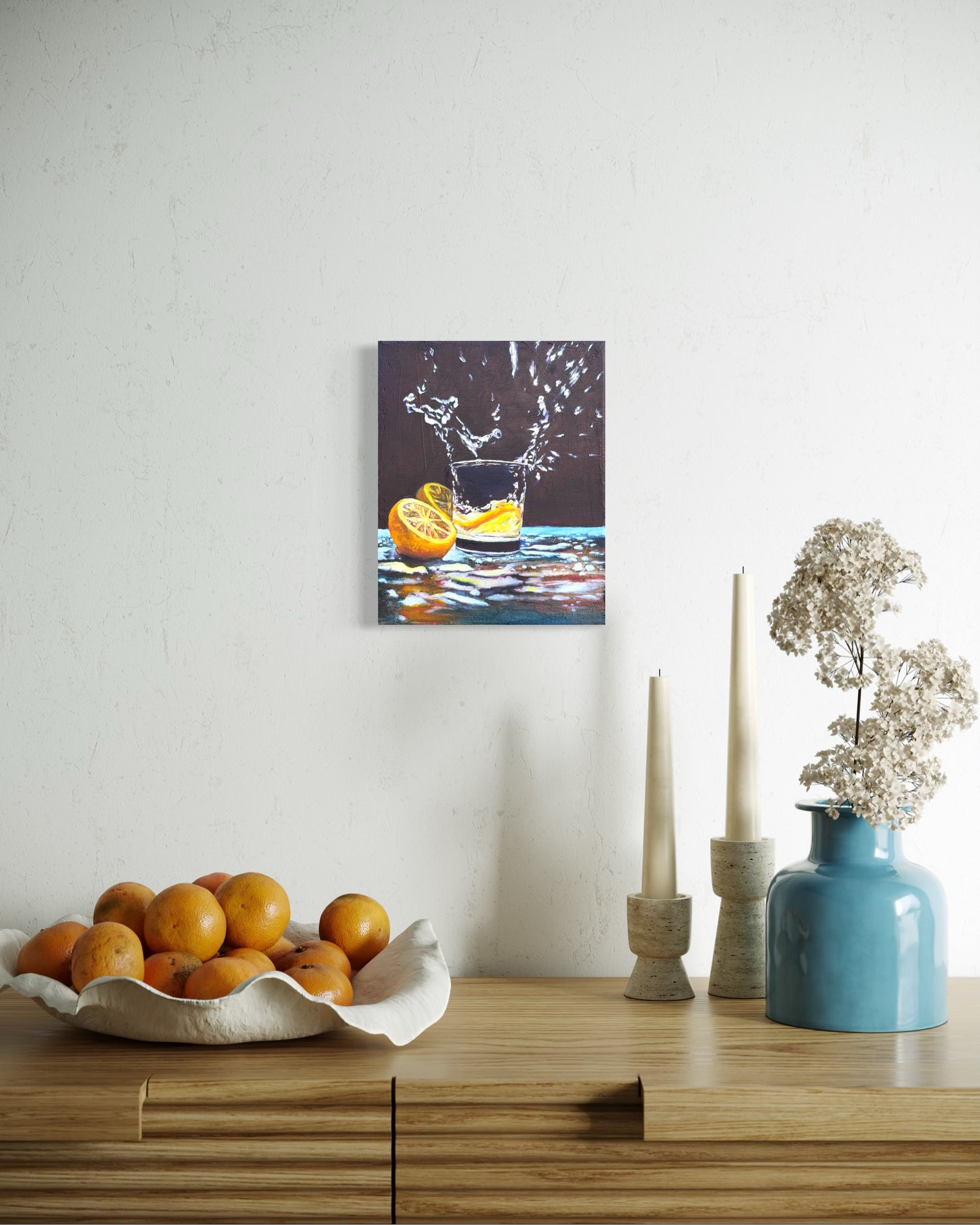 Decorative setup with a bowl of oranges, a vase with flowers, and an oil painting on a wall of lemon slices dropped in a glass of water.
