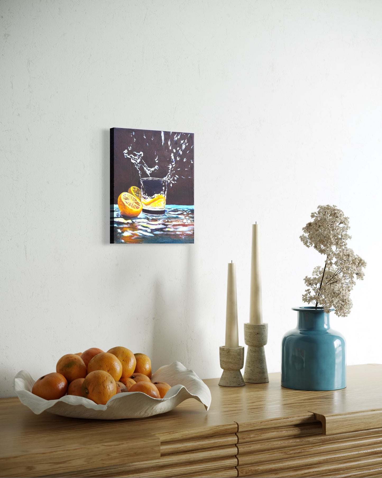 Canvas oil painting of a cup of water splashing out from an lemon slice dropped in and two halves beside the cup. Hung on a wall above a wooden table with decorative items.