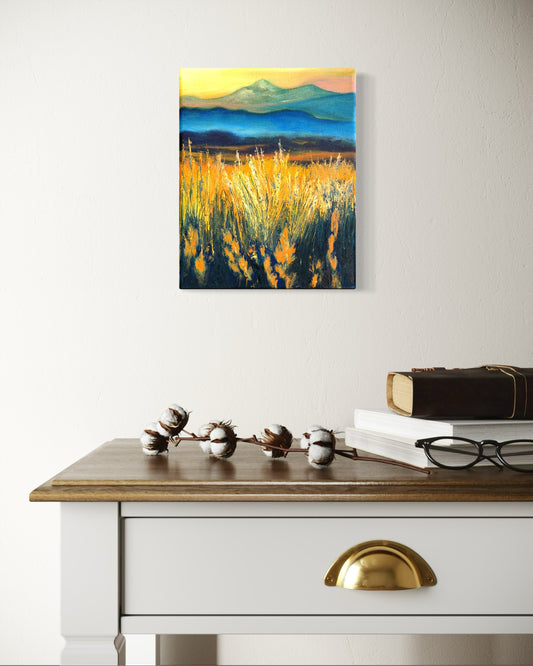 An oil painting of a faded mountain landscape with tall grass glowing from the sun on a wall above a desk.