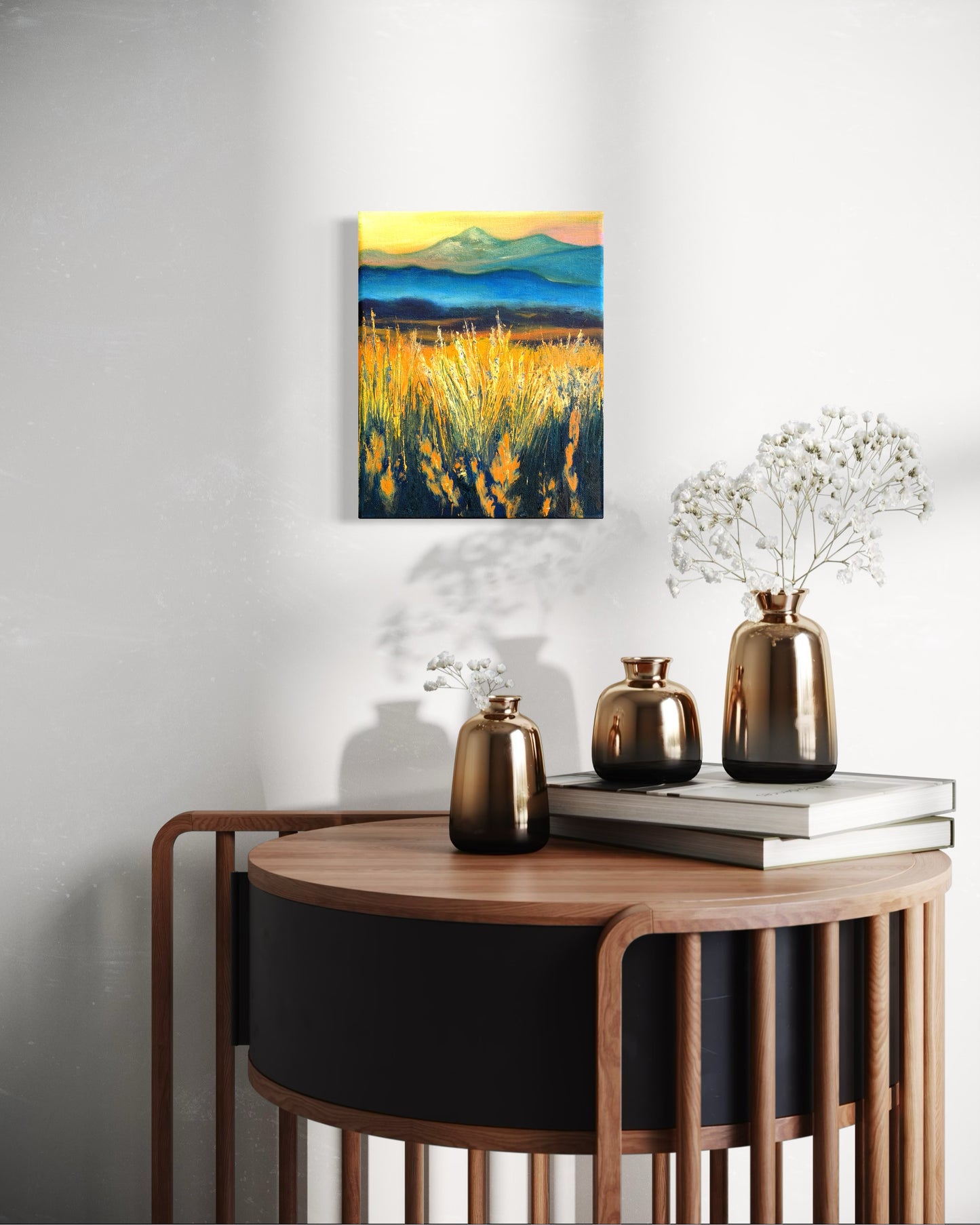 Decorative setting with a round wooden table and vases. An oil painting of a faded mountain landscape with tall grass glowing from the sun hangs on the wall.