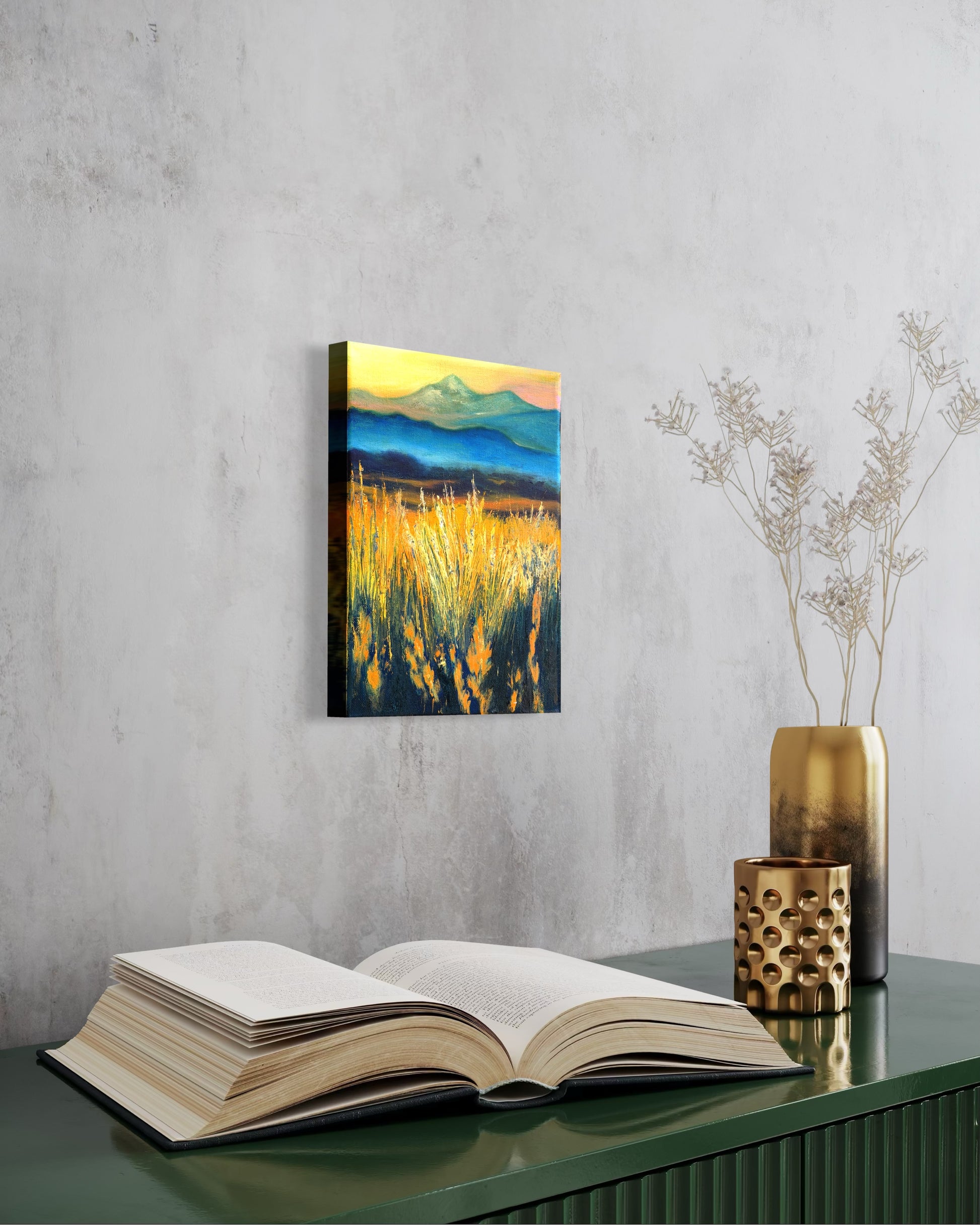 Canvas oil painting of a faded mountain landscape with tall sun glowing grass hanging on a wall above an open book.