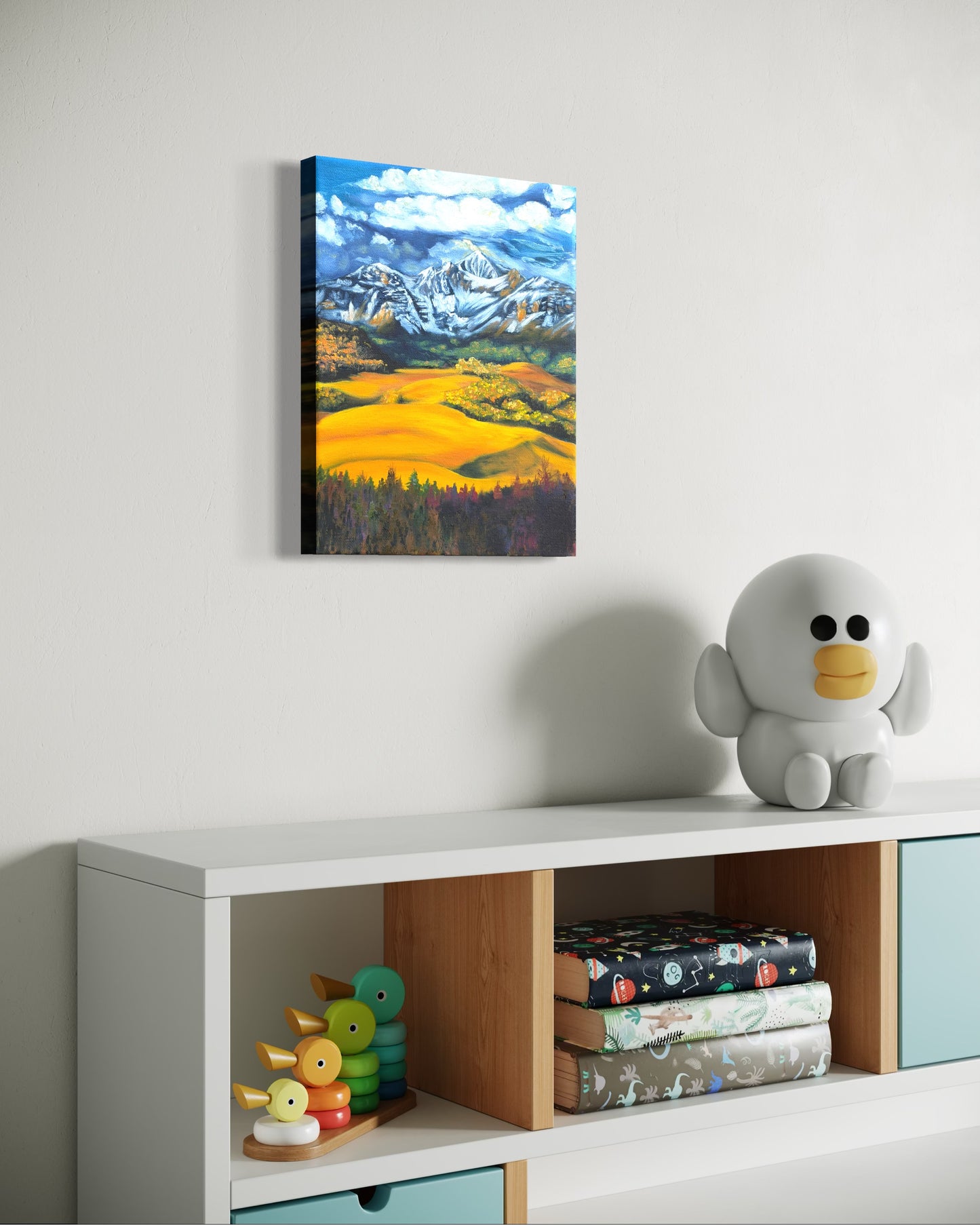 Oil painting of snow capped mountains and golden fields hanging on a wall above a shelf with toys and books.