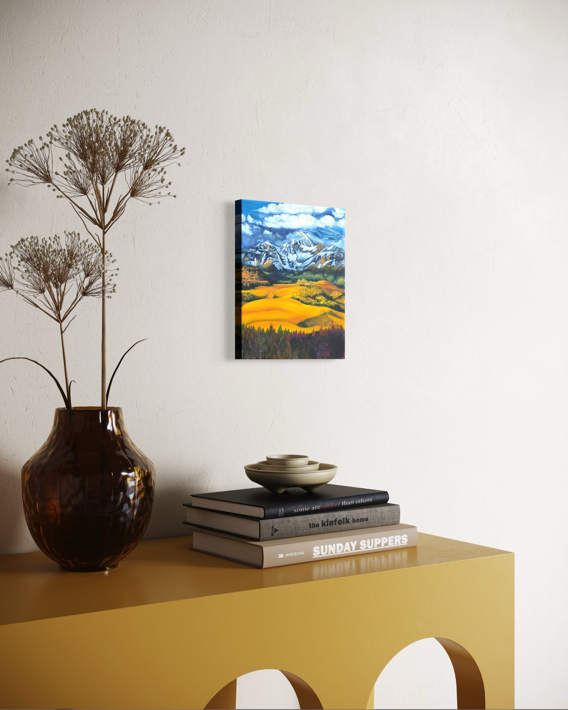 Decorative setup with a vase, books, and an oil painting of a mountain landscape with snow-capped peaks, green and golden valleys, and blue sky.