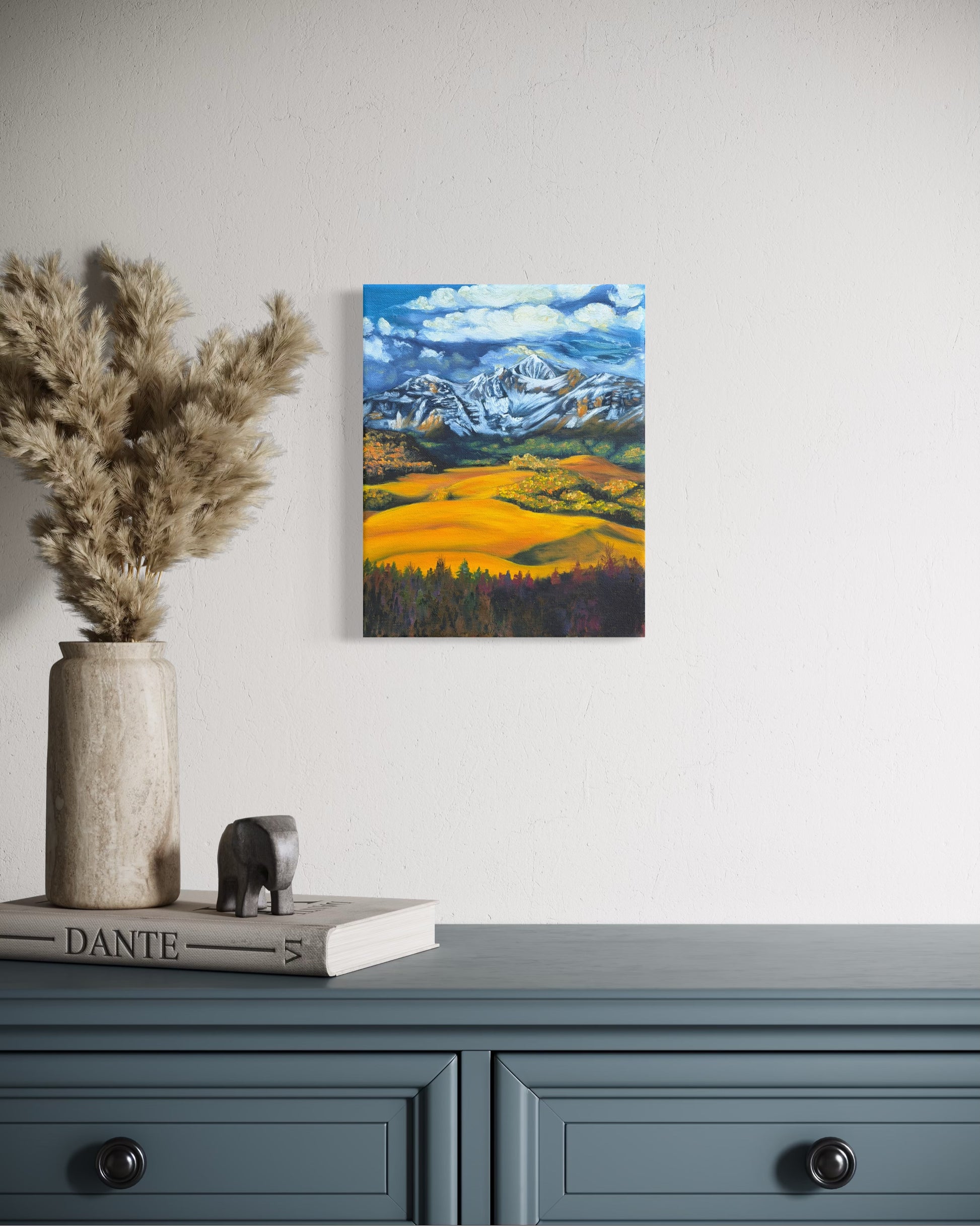 Painting of a mountain landscape with snow-capped peaks, green and golden valleys, and blue sky hanging on a wall above a blue cabinet with decorative items.