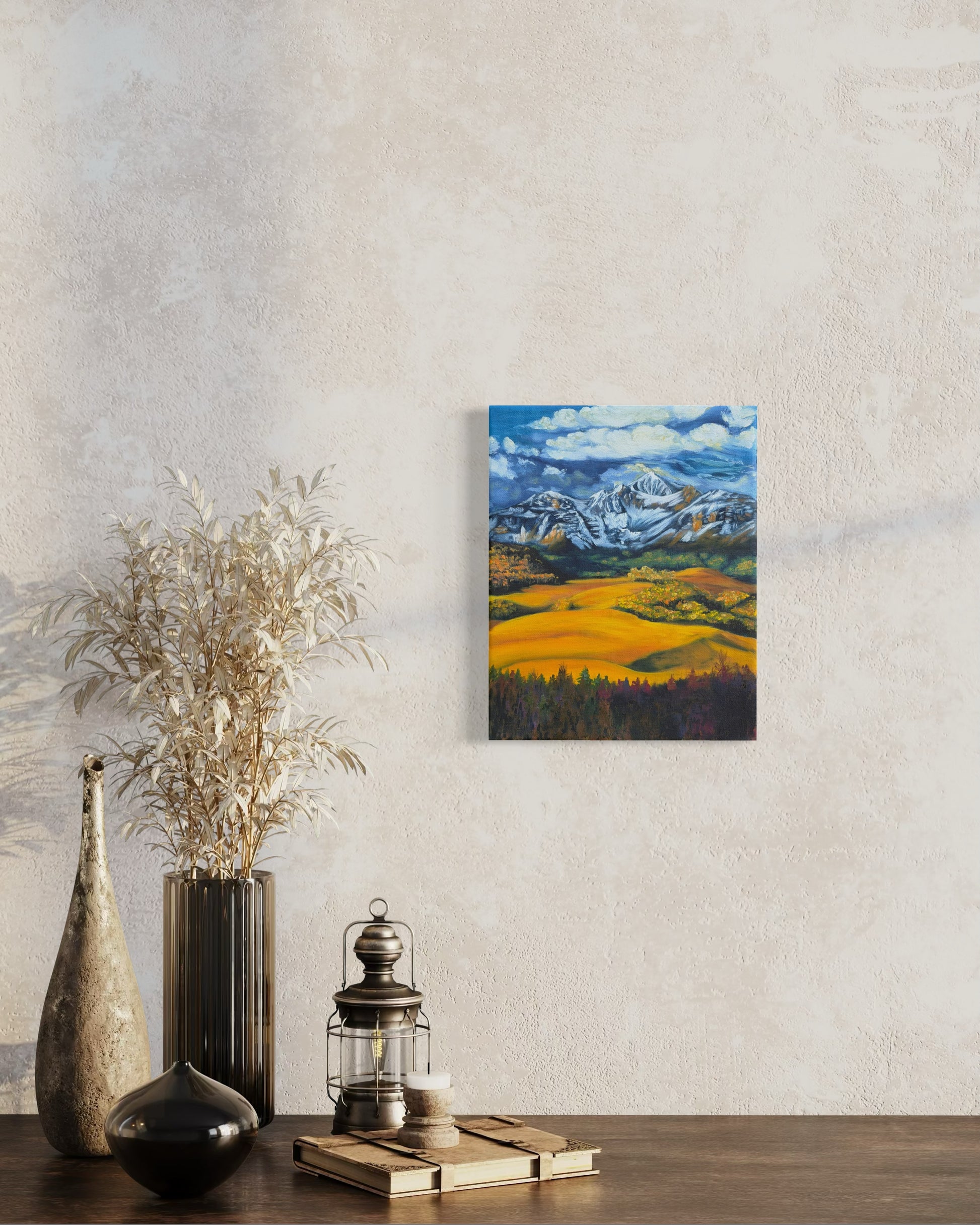 Painting of a mountain landscape with snow-capped peaks, green and golden valleys, and blue sky hanging on a wall above decorative items.