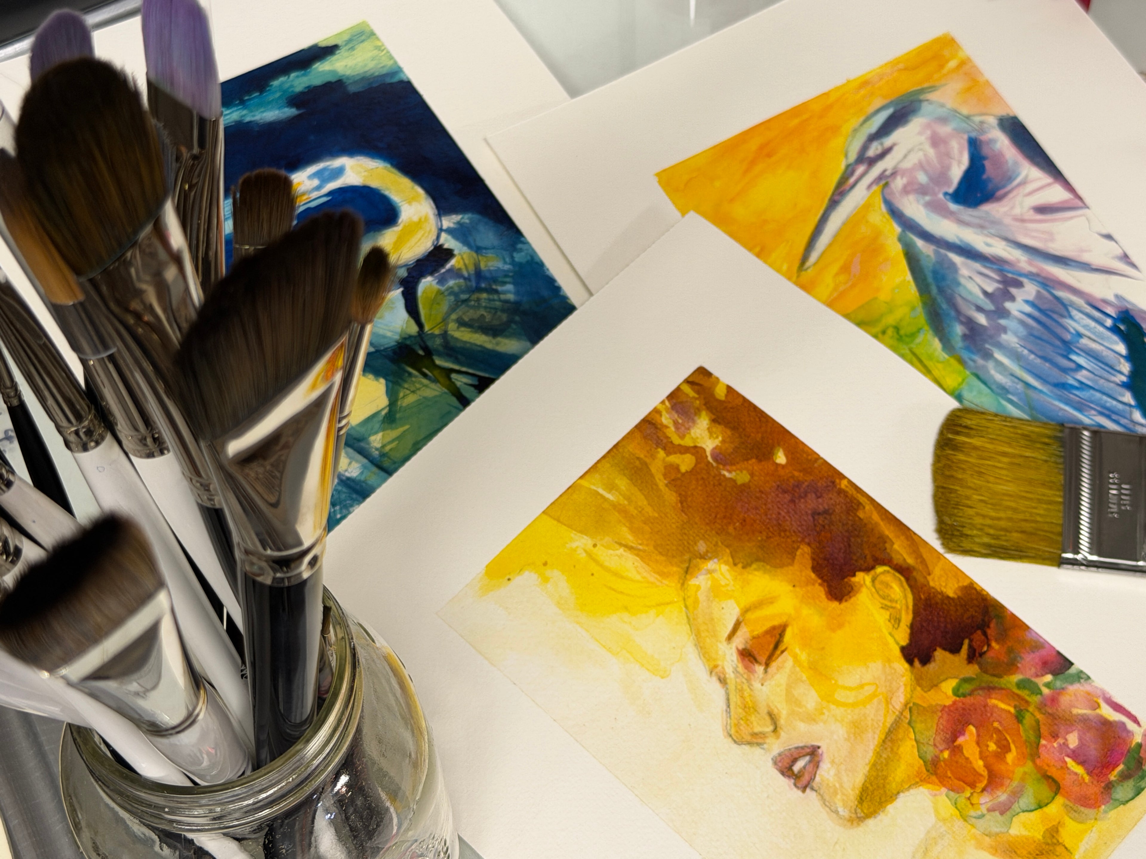 Closeup of three paintings flat on a desk with a cup of paint brushes.