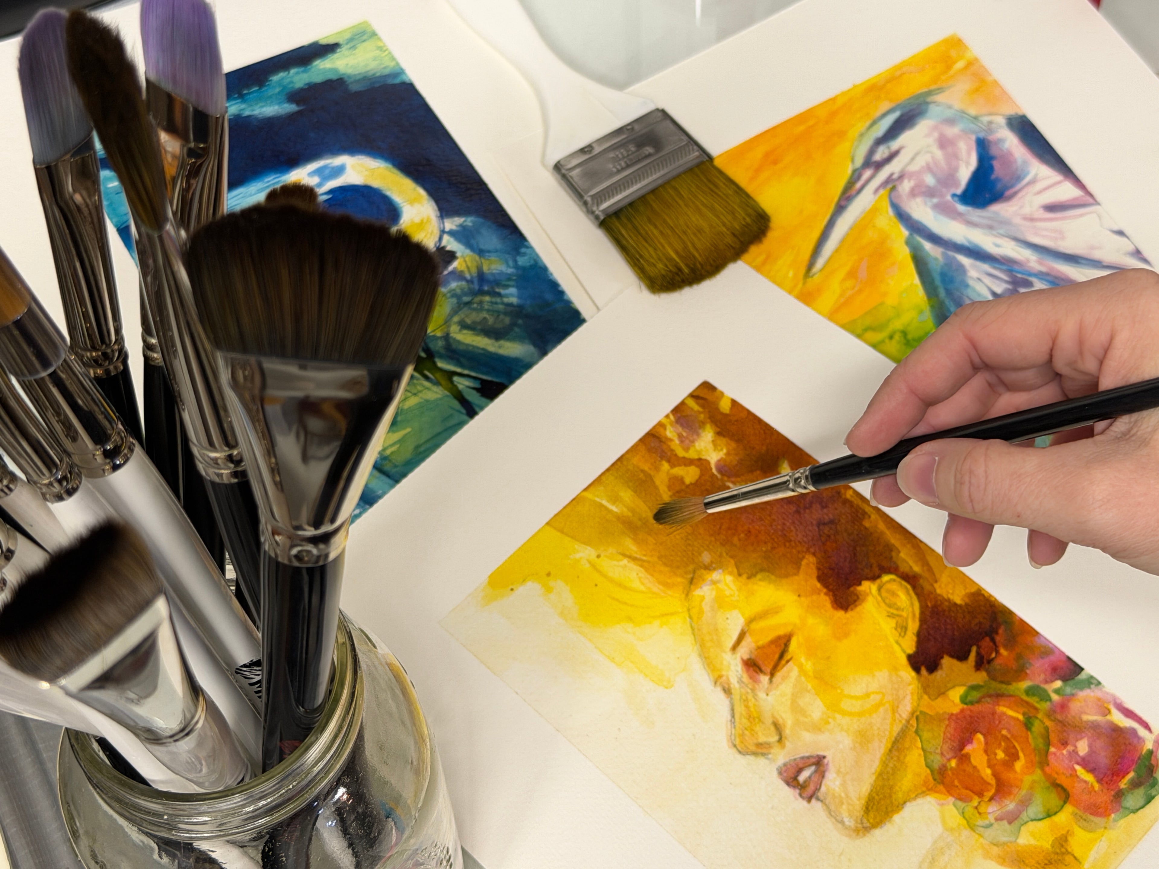 Close up of paint brushes surrounded by three paintings and a hand painting on a desk