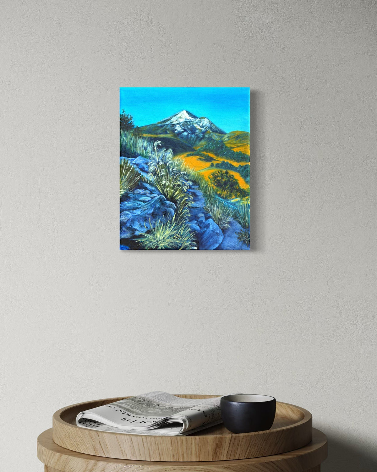 Canvas oil painting of a snow capped mountain landscape and a rocky path hanging on a wall above a wooden side table with a newspaper and mug.