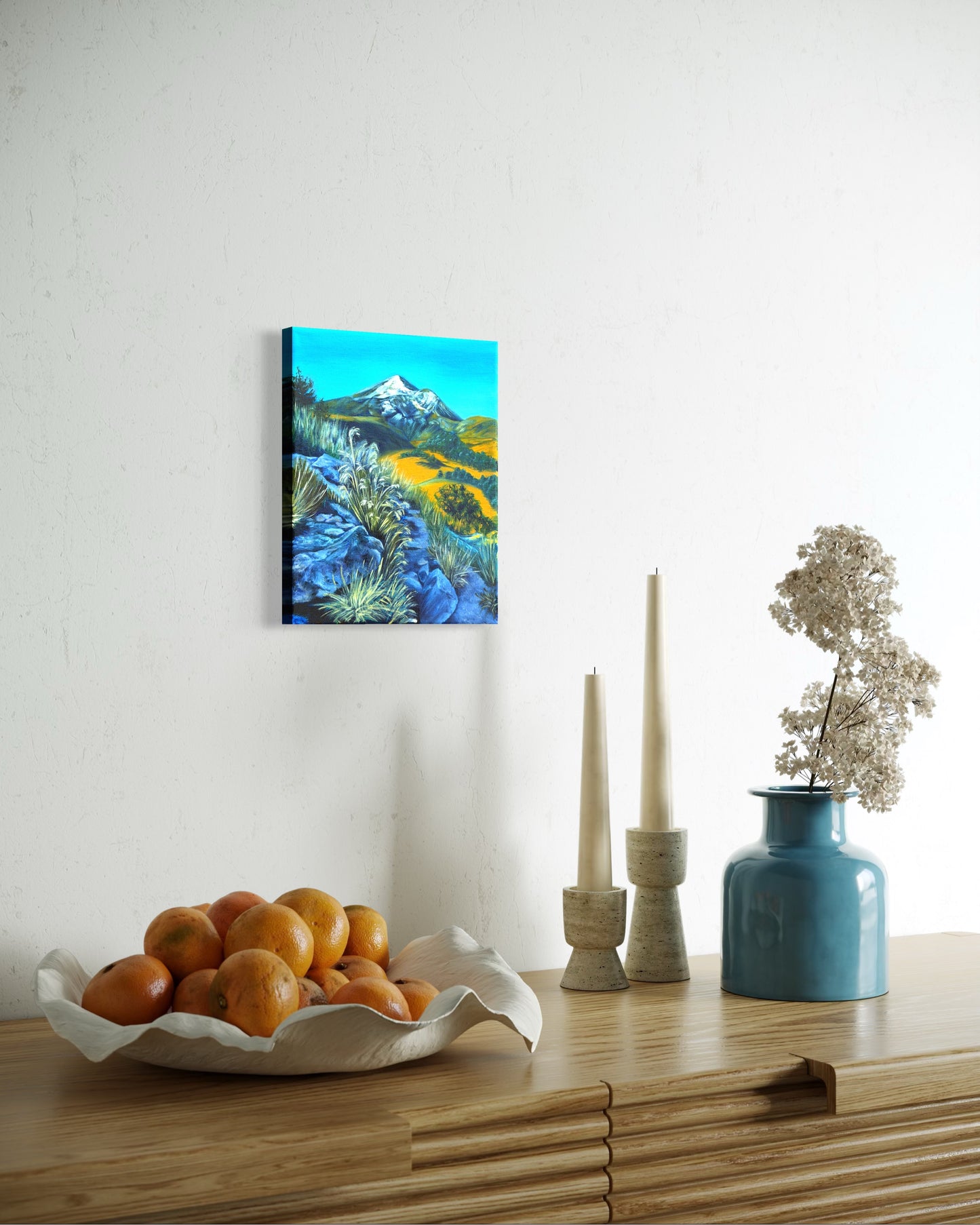 Decorative setup with an oil painting of a mountain landscape with a rocky path and greenery handing above a bowl of oranges, and vase on a wooden table.
