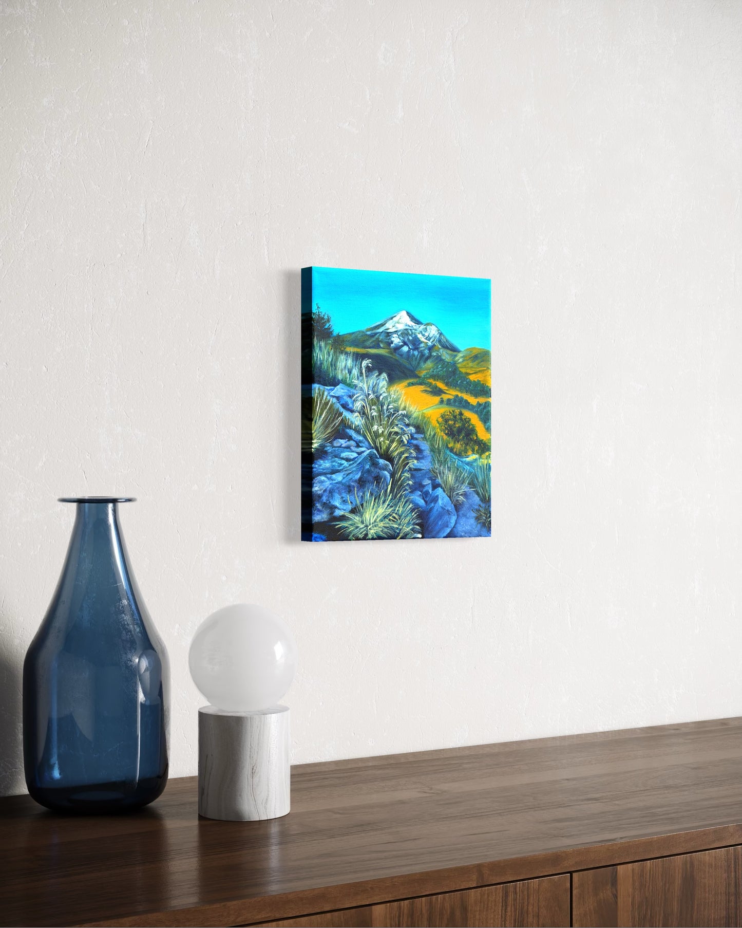 Oil painting of a mountain landscape and rocky path with greenery hanging on a wall above a wooden surface with decorative items.