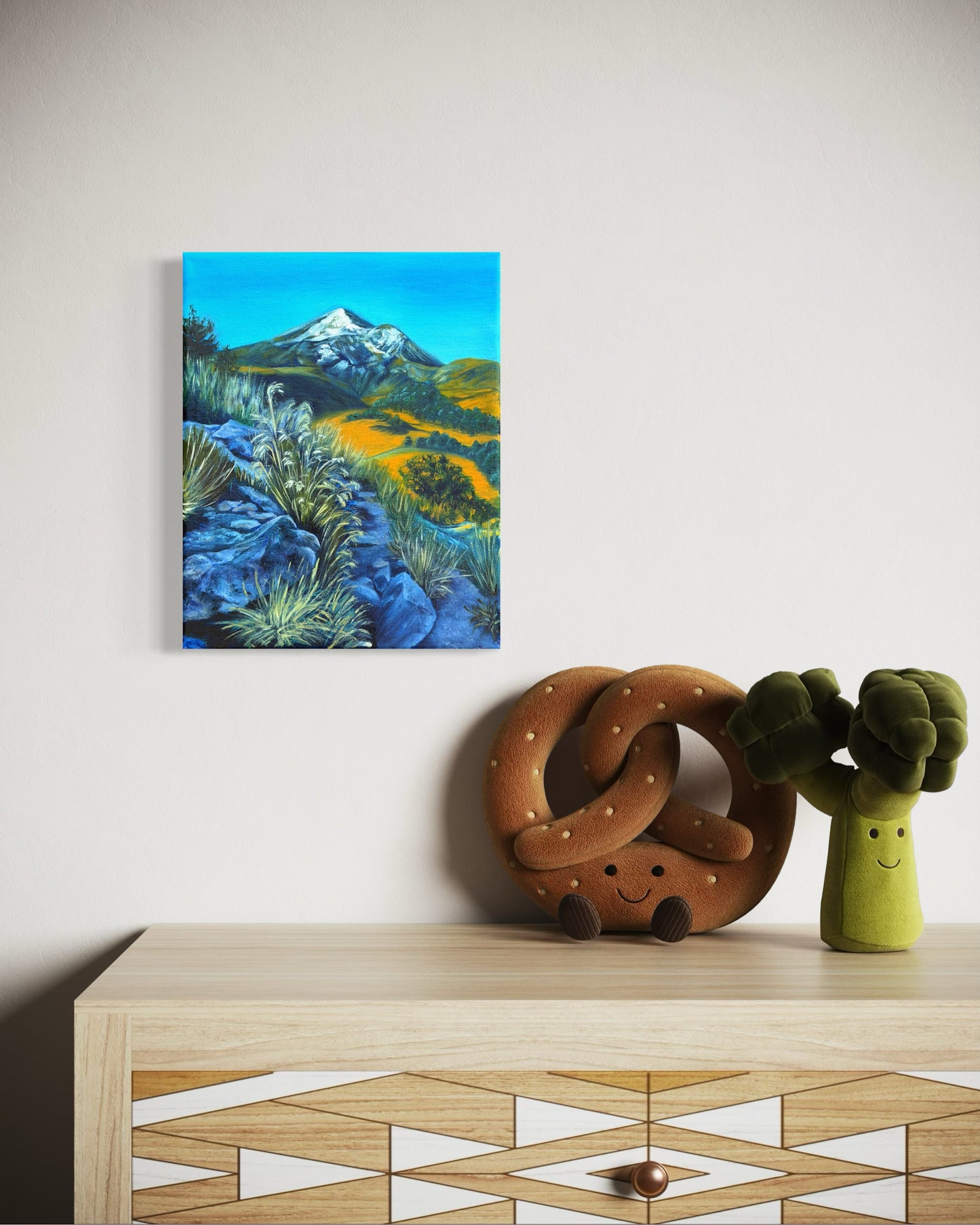 Oil painting artwork of a mountain landscape with rocky path and greenery hanging on a wall above a wooden cabinet with kid toy items.