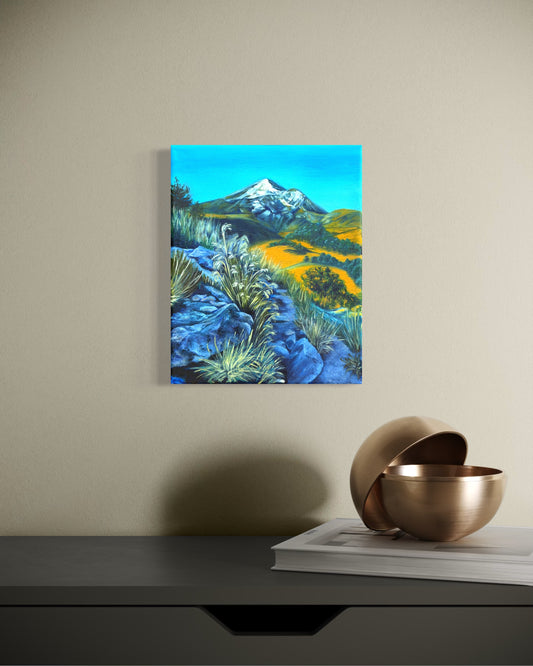 Canvas oil painting of a snow capped mountain landscape with a rocky path and greenery handing on a beige wall above a dark table with a metallic bowl.