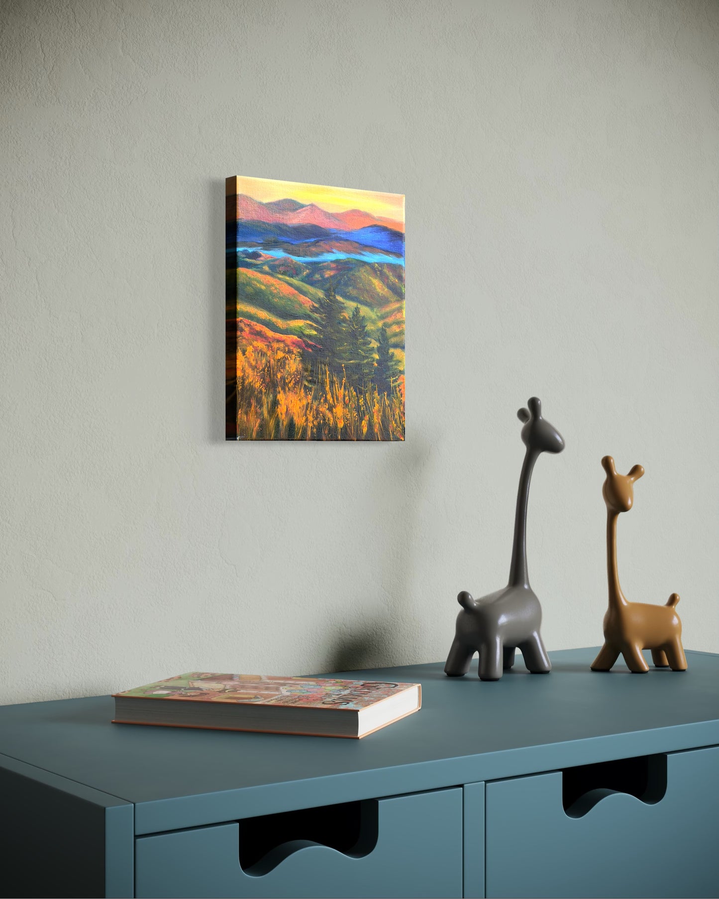 Oil painting of a landscape with rolling hills, trees, and a river in-between the mountains at sunset hanging on a wall above a blue cabinet with kid toy items.