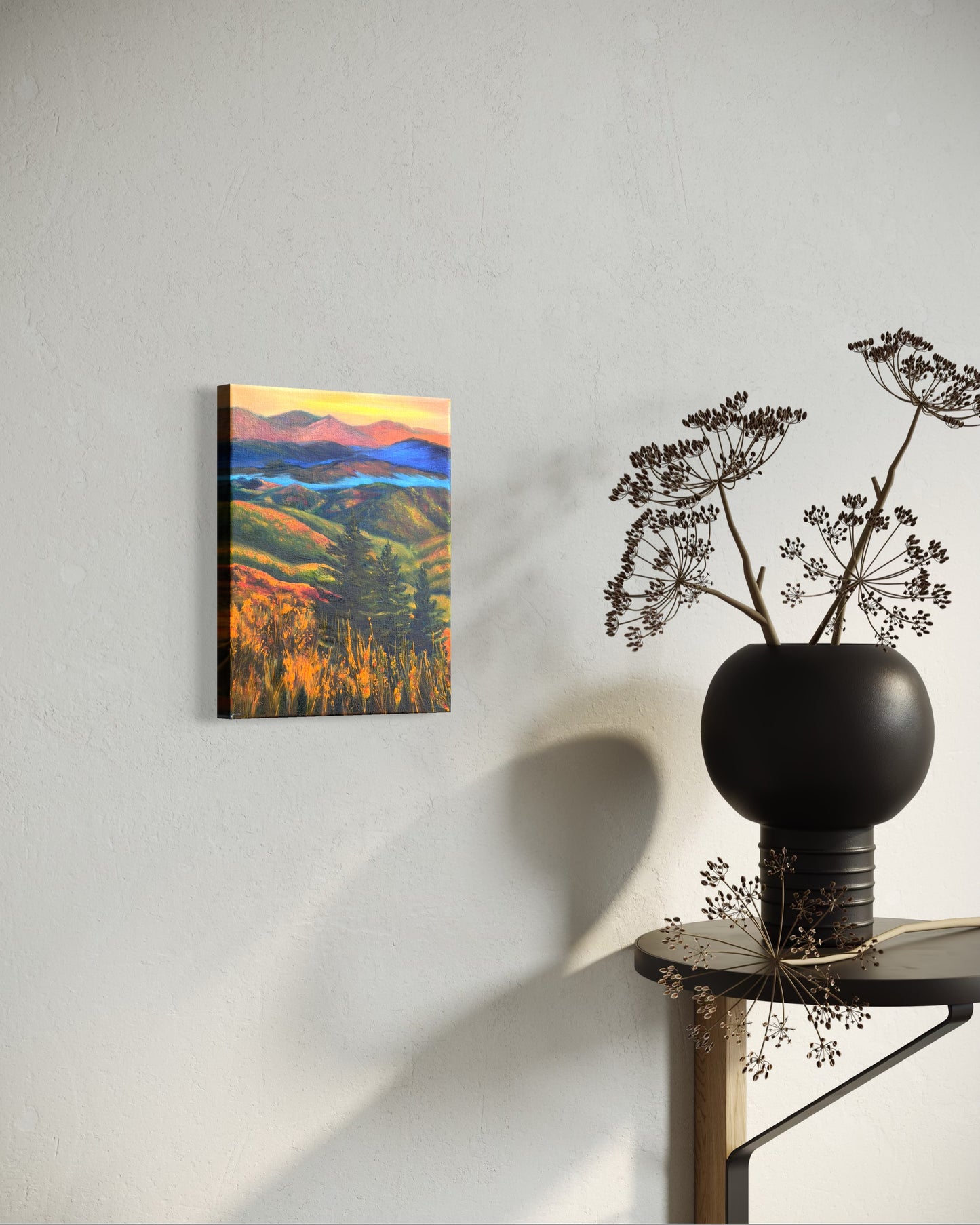 Oil painting of a landscape with rolling hills, trees, and a river in-between the mountains at sunset hanging on a wall next to a black vase with dried plants.