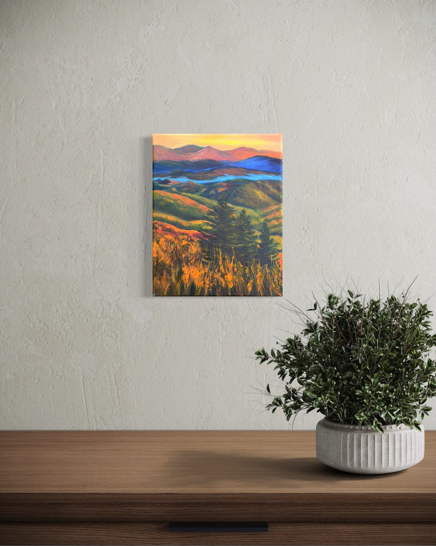Oil painting of a landscape with rolling hills, trees, and a river in-between the mountains at sunset hanging on a wall above a wooden surface with a plant.