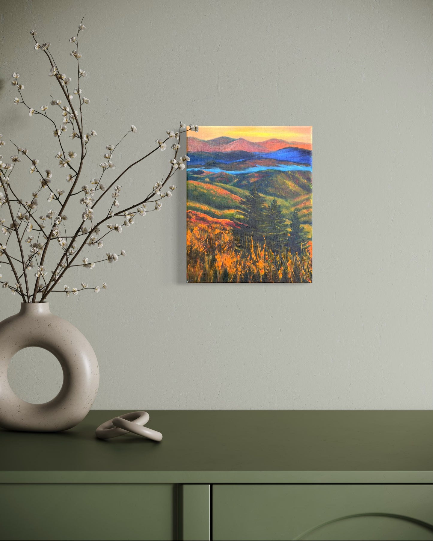 Oil painting of a landscape with rolling hills, trees, and a river in-between the mountains at sunset hanging on a wall above a green cabinet with decorative elements.