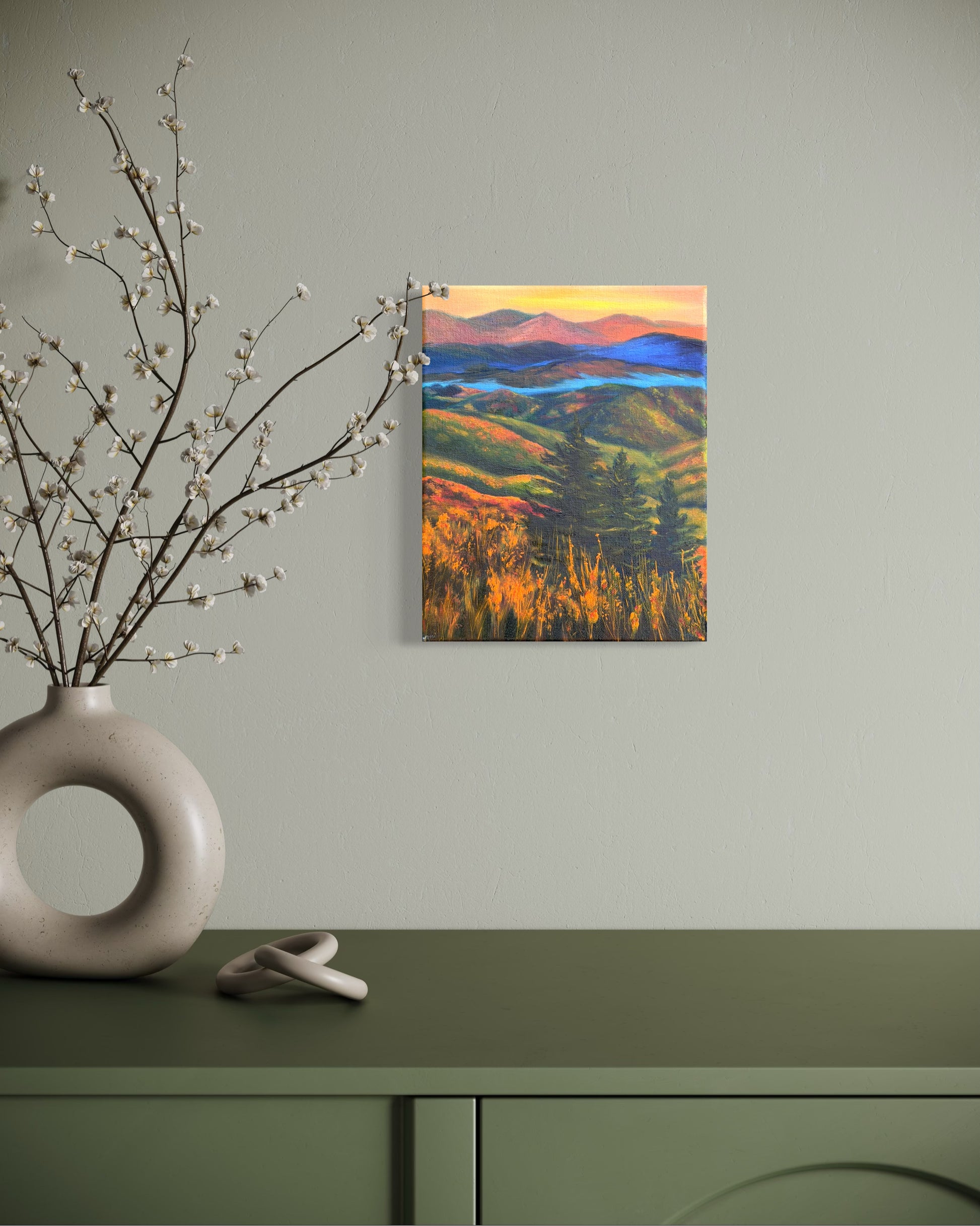 Oil painting of a landscape with rolling hills, trees, and a river in-between the mountains at sunset hanging on a wall above a green cabinet with decorative elements.